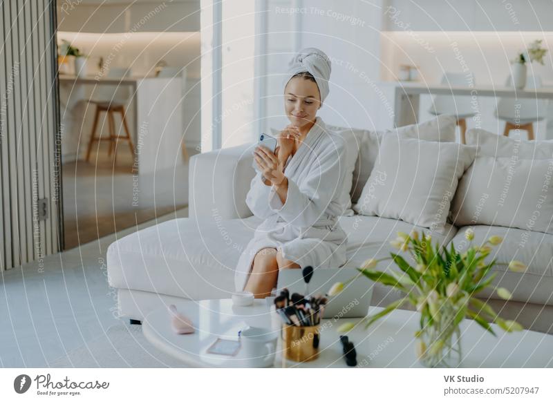 Young pretty young woman looks at modern smartphone camera takes selfie has well cared healthy skin applies cream wears bathrobe and towel poses on sofa does cosmetic procedures for complexion