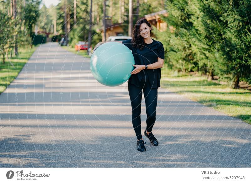 Fitness woman exercises with fitness ball outdoor walks on road near forest has healthy lifestyle dressed in black active wear, has happy expression, stays in good physical shape. Fitness concept