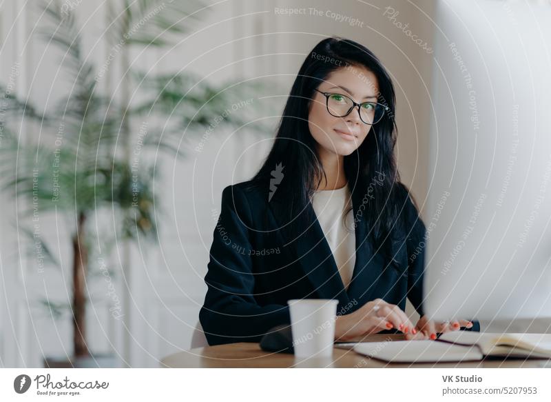 Serious successful female office worker in formal outfit, wears eyeglasses, busy with online work, poses at desktop, works on computer, reads business new or watches tutorial, enjoys working process.