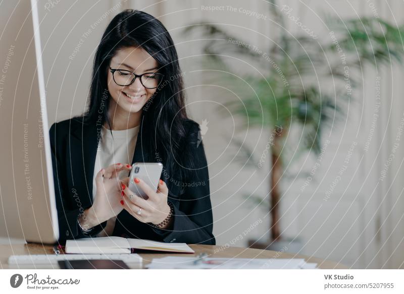 Beautiful smiling female freelancer works remotely, concentrated in smartphone, sends feedback on received message, sits in coworking space, dressed formally, works on computer, surfs internet