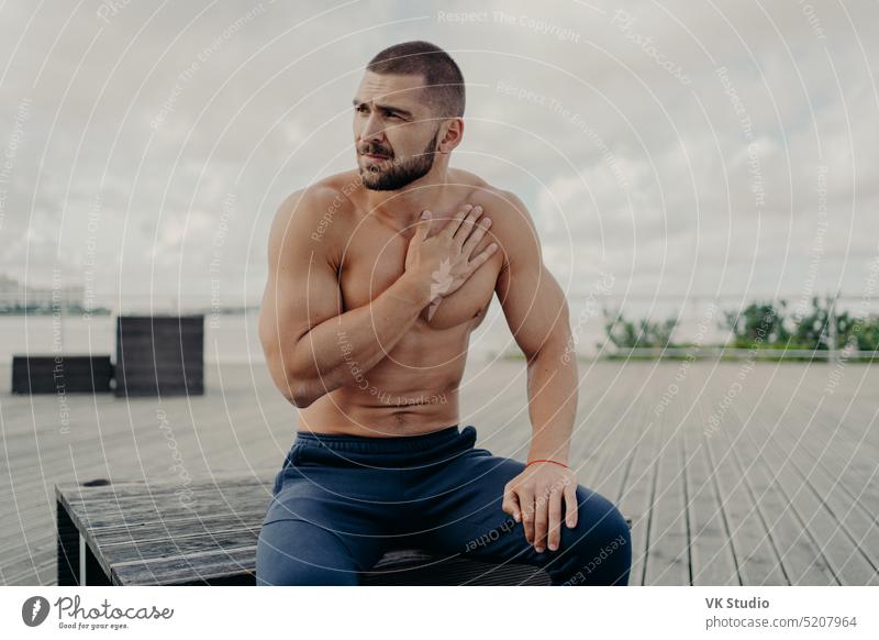 Thoughtful bearded man has rest after intensive cardio training, keeps hand on chest and has shortage of breathing, sits outdoor, concentrated aside, poses topless. Sport and healthy lifestyle