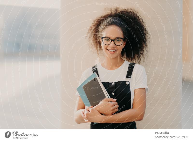 Adorable curly haired female student wears white casual t shirt and overalls, holds notepad or textbook, looks through transparent spectacles, stands against white background. People and lifestyle