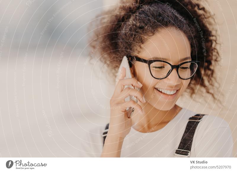 Close up portrait of cheerful woman with crisp hair, satisfied with tariffs for telephone call, focused down, holds modern smart phone, phones to best friend, discusses latest news. Technology concept