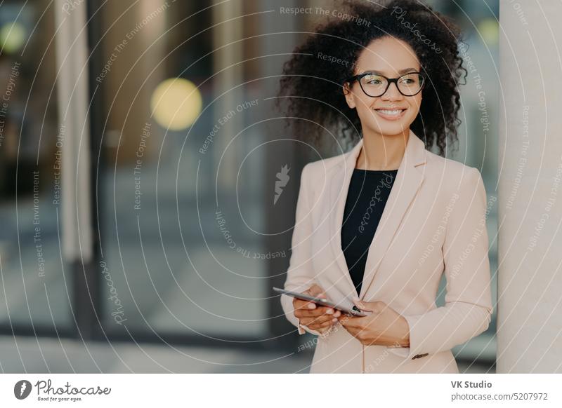 Positive successful woman entrepreneur with Afro hair holds digital tablet, stands outdoor near office building, wears formal clothes, looks away, waits for colleague to have dinner during break