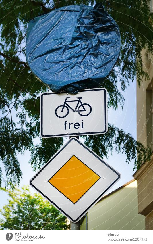 Traffic sign right of way road, additional sign bicycle traffic free and a round traffic sign covered with blue plastic tarpaulin on a metal pipe under a tree and next to a residential house