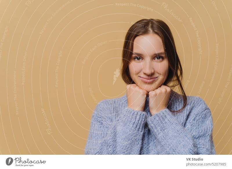 Young European woman with minimal makeup, dark hair, dressed in winter sweater, keeps hands under chin, has charming smile, stands against brown background, copy space for your advertising content