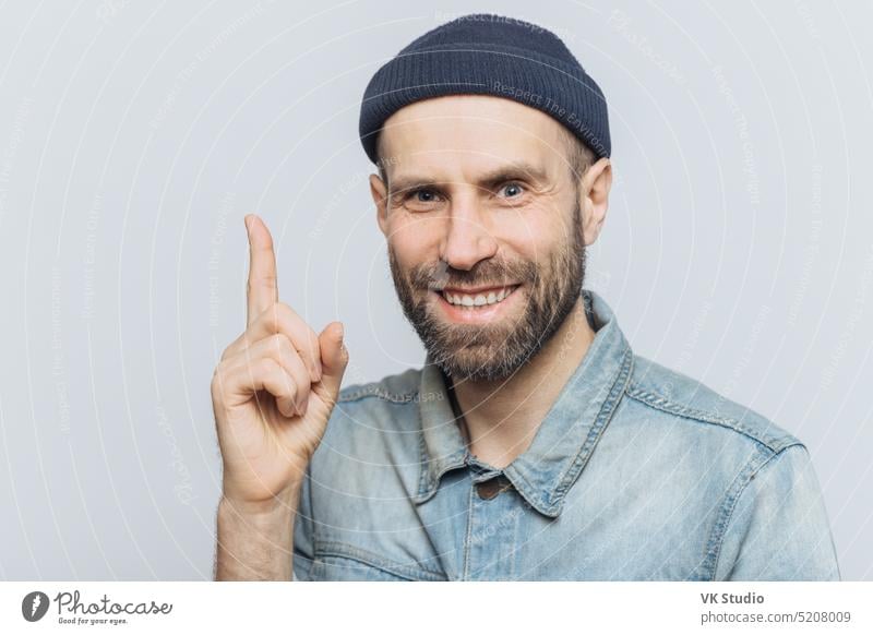 Attractive middle aged male raises fore finger as gets good idea in mind, wears fashionable clothing, has pleasant smile. Businessman keeps finger up, happy to remember new successful investing plan
