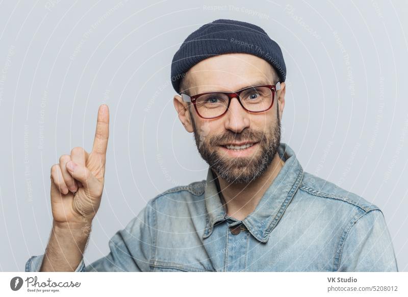 People, emotions and reaction concept. Handsome bearded guy in trendy eyewear, has pleased expression, raises fore finger as remembers something positive, isolated over grey studio background