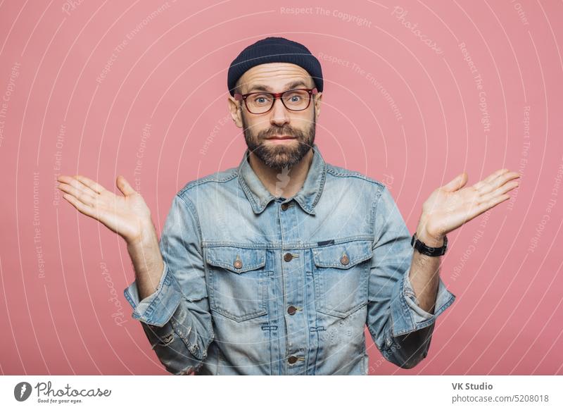 Photo of attractive bearded male wears fashionable clothing, shrugs shoulders in bewilderment, makes serious decision, isolated over pink background. Confused hesitant unshaven stylish man