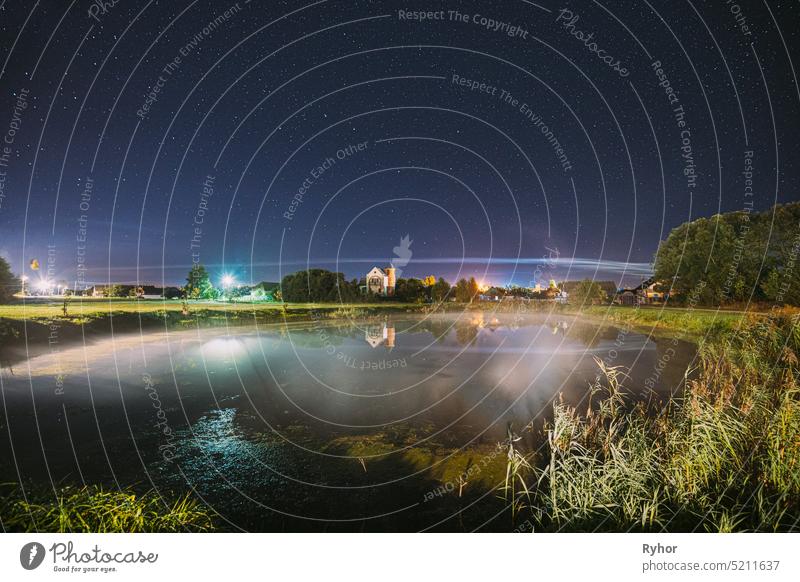 Night Clouds Over River Lake Near Houses In Village. Night Starry Sky Above Lake River With Bright Stars And Meteoric Track Trail. Glowing Stars Above Summer Nature