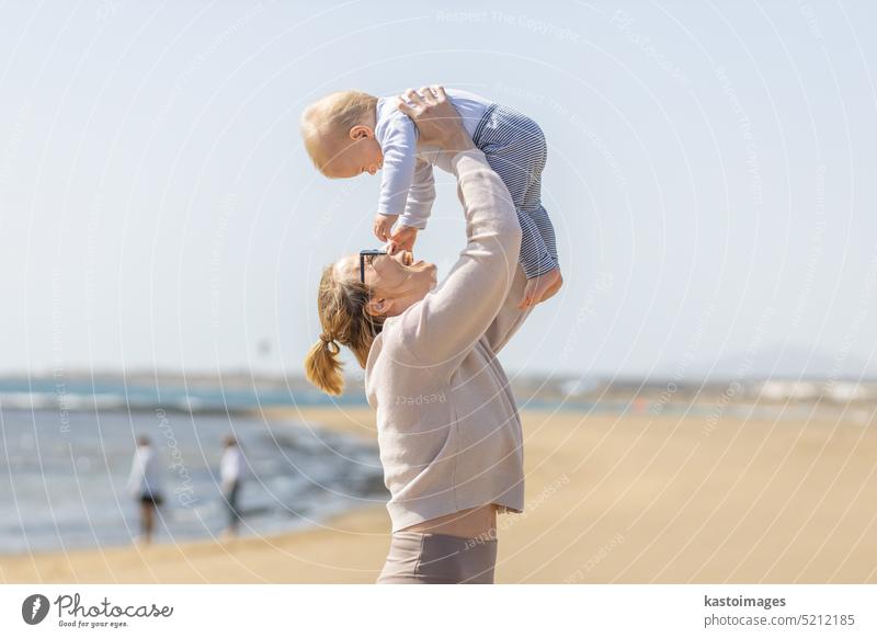 Mother enjoying summer vacations holding, playing and lifting his infant baby boy son high in the air on sandy beach on Lanzarote island, Spain. Family travel and vacations concept