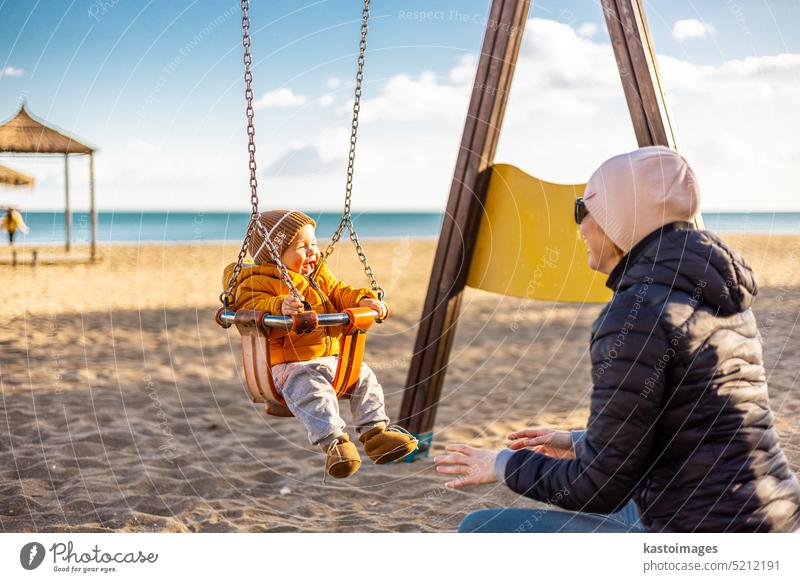 Mother pushing her infant baby boy child on a swing on sandy beach playground outdoors on nice sunny cold winter day in Malaga, Spain. happy son family kid