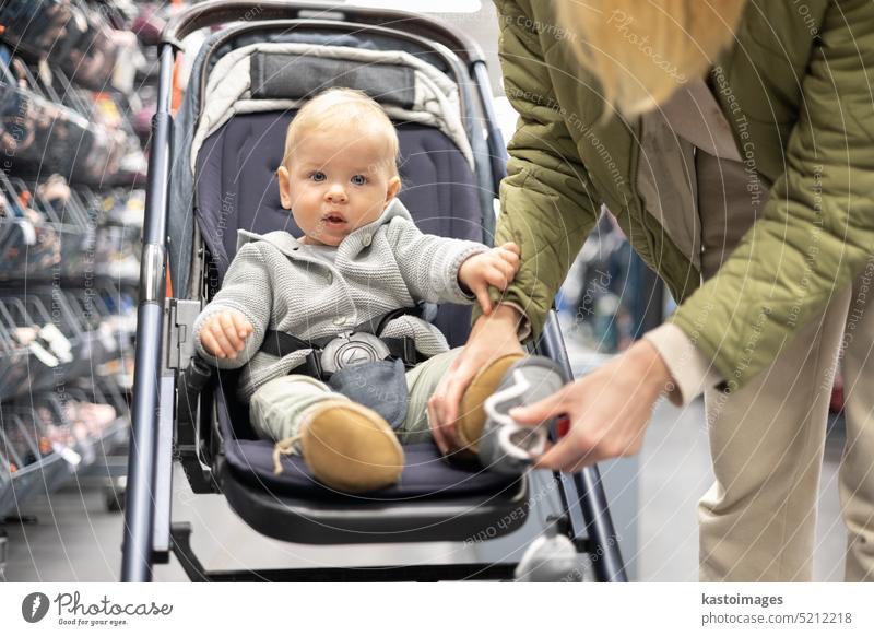 Casualy dressed mother choosing sporty shoes and clothes products in sports department of supermarket store with her infant baby boy child in stroller. woman