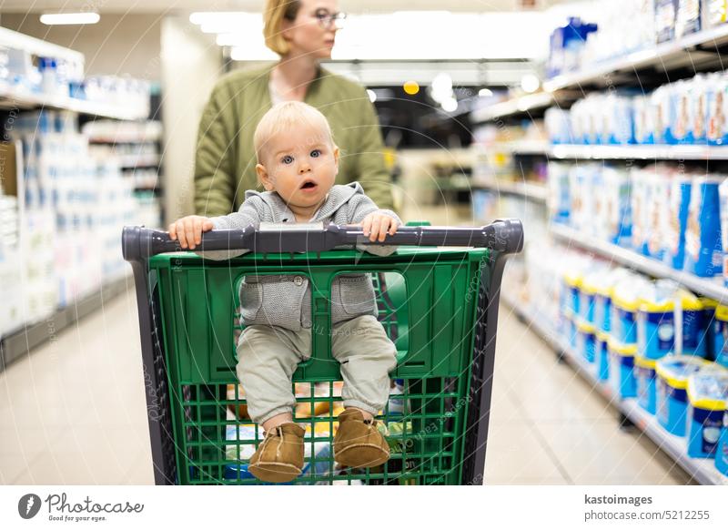 Mother pushing shopping cart with her infant baby boy child down department aisle in supermarket grocery store. Shopping with kids concept. woman mom young