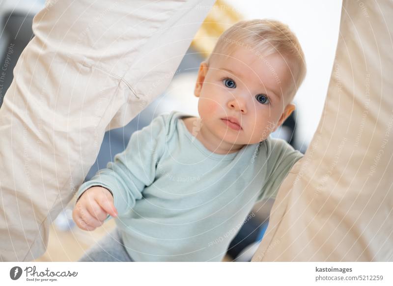 Portrait of adorable curious infant baby boy child taking first steps holding to father's pants at home. Cute baby boy learning to walk. portrait standing small