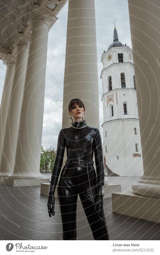 Next to Vilnius cathedral and bell tower, a gorgeous brunette girl in black latex is posing for a model test. A fashion model is wearing a latex catsuit and being all pretty. Her outfit is outstanding. And so does she.