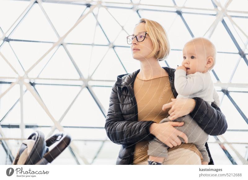 Mother carying his infant baby boy child, pushing stroller at airport departure terminal waiting at boarding gates to board an airplane. Family travel with baby concept.