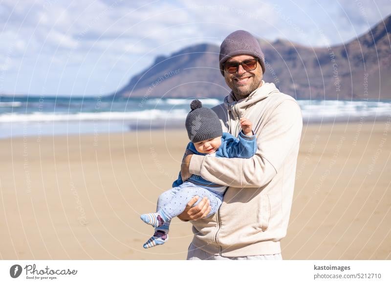 Father enjoying pure nature holding and playing with his infant baby boy son in on windy sandy beach of Famara, Lanzarote island, Spain. Family travel and parenting concept.