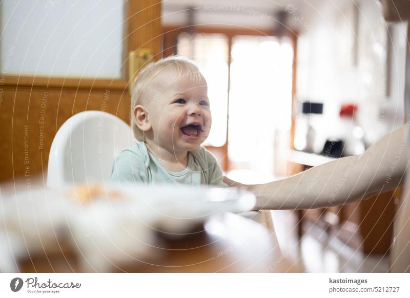 Adorable cheerful happy infant baby boy child smiling while sitting in high chair at the dining table in kitchen at home beeing spoon fed by his mother food
