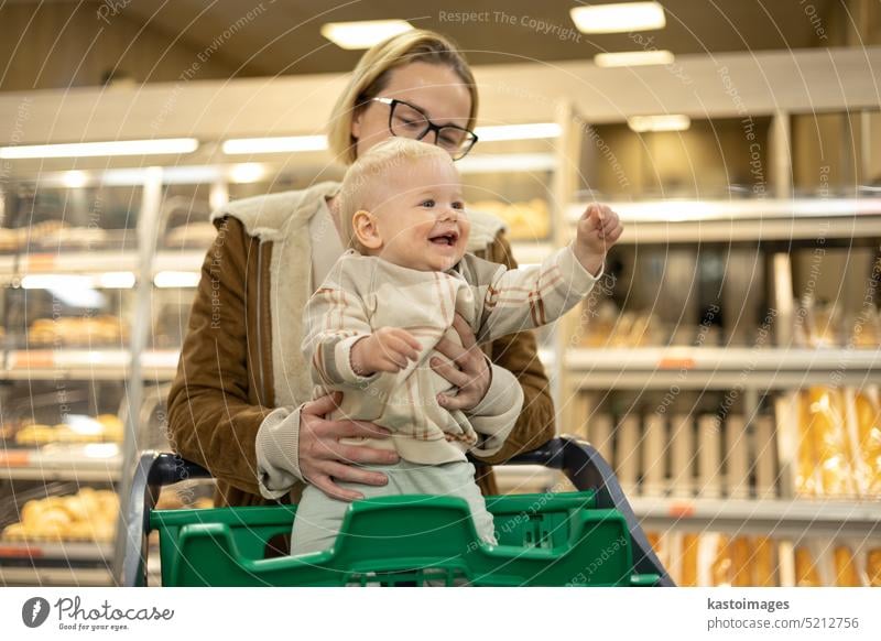 Mother shopping with her infant baby boy, holding the child while stacking products at the cash register in supermarket grocery store. woman mom kid young