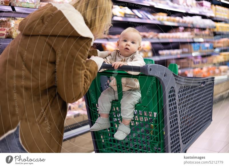 Mother shopping with her infant baby boy, holding the child while stacking products at the cash register in supermarket grocery store. woman mom kid young