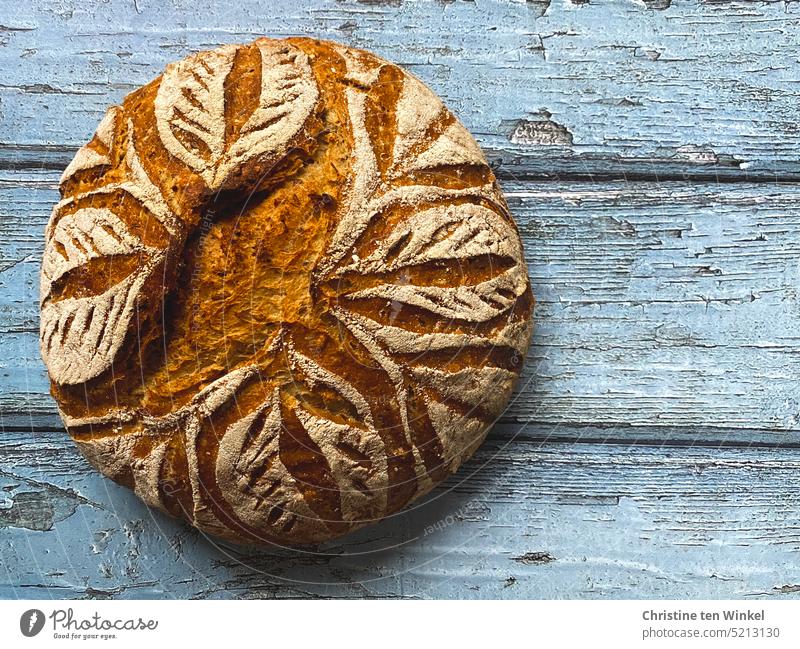 An oven warm fragrant bread loaf with leaf pattern lies on light blue background our daily bread homemade To enjoy Appetite Bird's-eye view Self-made