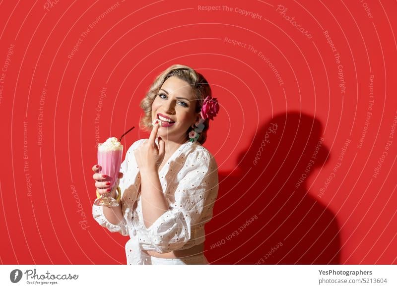 Woman drinking a milkshake against a red background. Retro style woman portrait 50s beauty blonde blue eyes color concept cream curly curvy cute dress enjoy