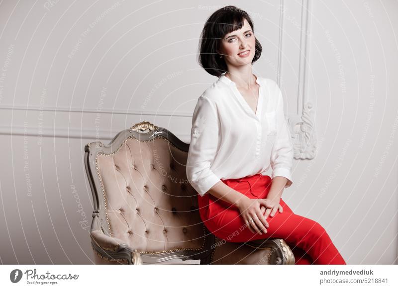 Studio portrait of attractive young woman with dark hair in white shirt and red pants sitting on classic chair on plain white background.Bossy female, job interview concept,confident independent lady