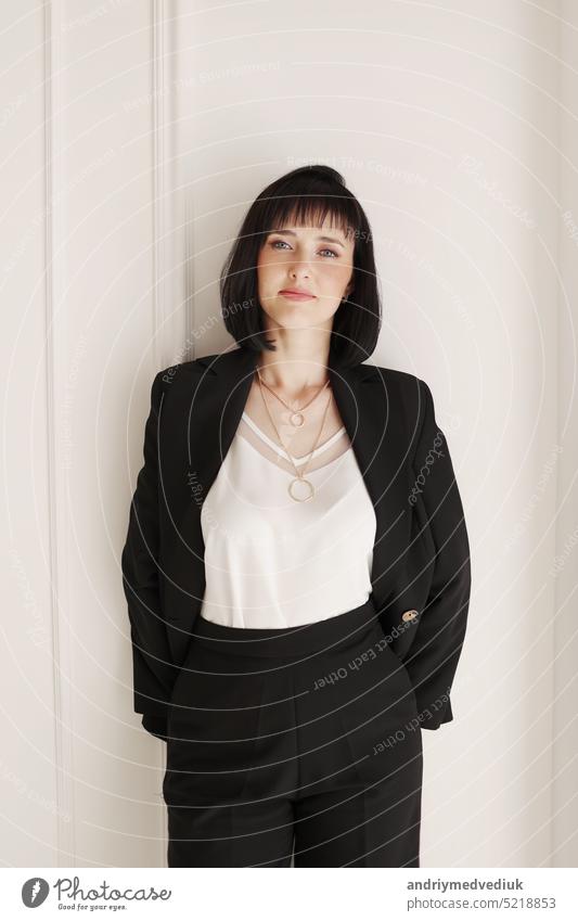 Confident young brunette business woman looking at camera on white wall background. Studio portrait of successful friendly female with short hair in black suit and white blouse with jacket on shoulder