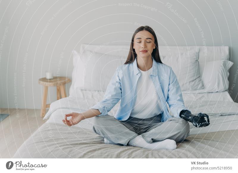 Disabled girl with bionic arm prosthesis practice yoga, meditating in lotus pose on bed at home disabled meditate breathe wellness calmness hand padmasana