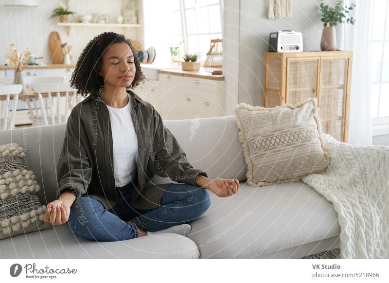 Calm mixed race girl does yoga at home, sitting in lotus pose on sofa. Meditation, healthy lifestyle meditate wellness calm breathe couch relax rest living room