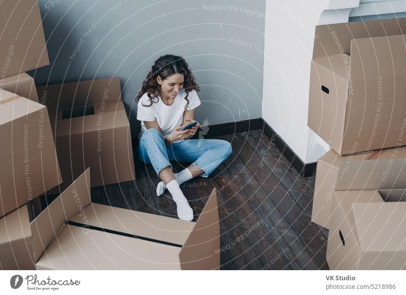 Modern girl sits with boxes uses smartphone app, choosing moving company and mover for relocation female cardboard box carton new home hispanic floor removal