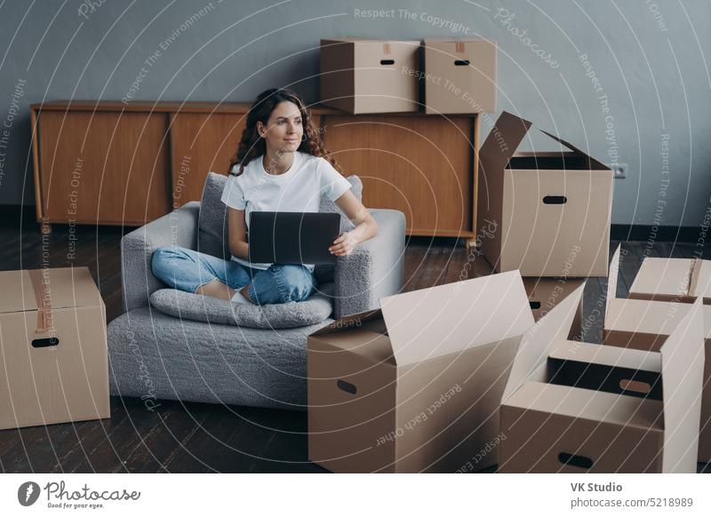 Happy girl sitting at laptop with cardboard boxes, dreaming about new first own home, relocation female new home carton removal armchair moving work spanish