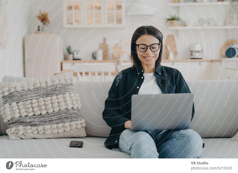 Smiling female working on laptop, shopping in online store, sitting on cozy couch at home. Ecommerce e commerce elearning education purchase remote girl chat