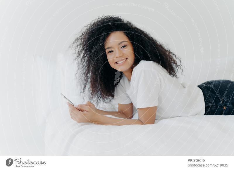 Pretty cheerful woman with Afro haircut, enjoys chatting and networking, lies on comfortable bed, looks photos, has relaxed face expression, isolated over white background. Online communication