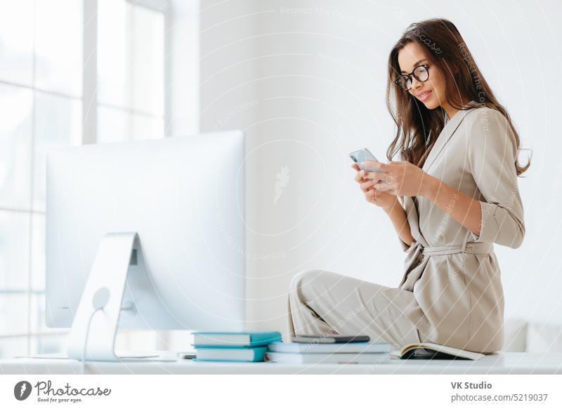 Horizontal shot of beautiful young woman in elegant clothes, has gentle smile, checks newsfeed via modern smartphone, has rest after hard work, poses in office interior at desktop, white walls