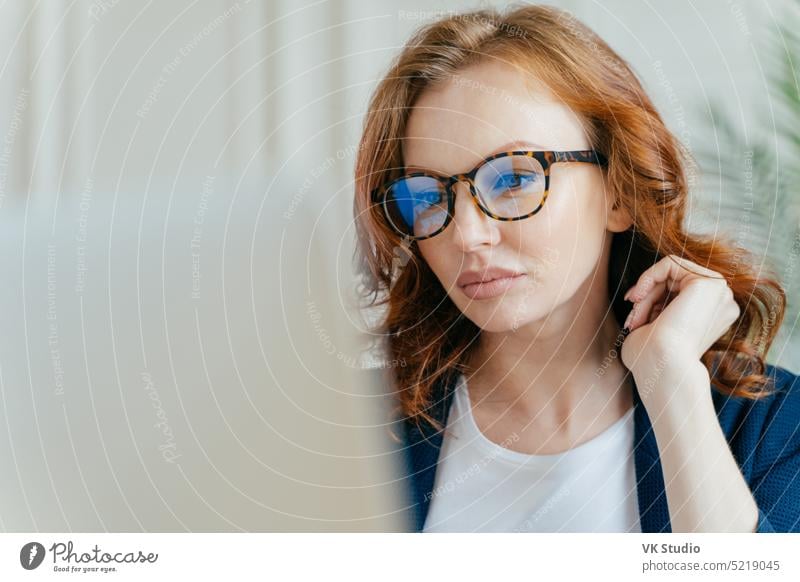 Close up shot of focused female worker completes successful project, makes remote job, focused in monitor of laptop computer, wears optical glasses for vision corrrection, has ginger curly hair
