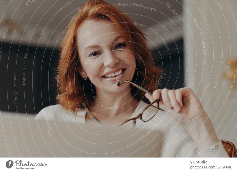 Cheerful redhead female web designer works on freelance, poses at opened laptop computer, holds glasses, shares content in social networks, smiles gently, poses at home. People and technology