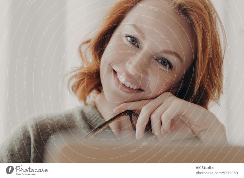 Close up shot of happy beautiful redhead woman tilts head, smiles positively, has white perfect teeth, makeup, sits in front of opened laptop computer, searches necessary information, uses free wifi