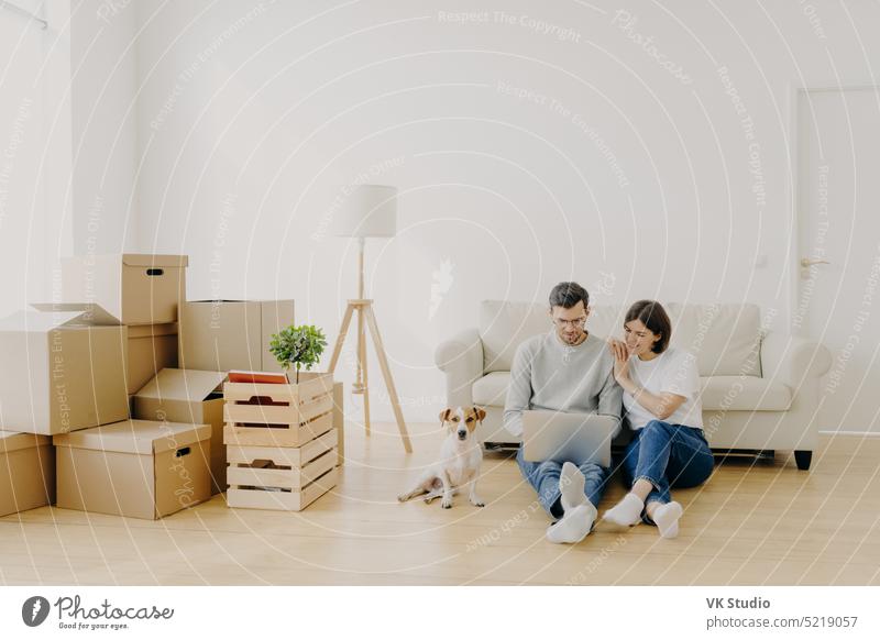 Affectionate married couple move in new home, relax on floor after unpacking things use laptop computer for searching home decoration ideas in internet their pet sits near, pose in purchased apartment