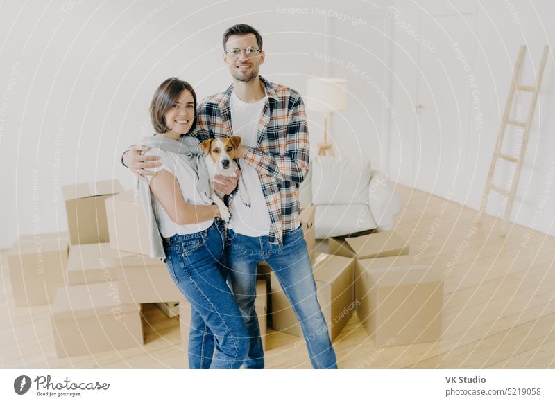 Happy woman and man tenants or renters of flat pose in own house, cuddle and pose with little dog, have glad expressions, start living in new bought apartment. Family and relocation concept.