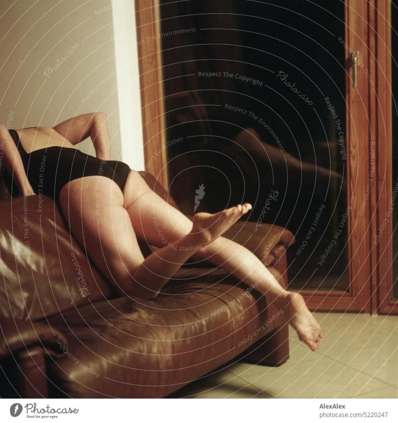 slim, athletic woman in black bodysuit kneels with one leg on a couch over the backrest and holds her legs in the air in front of a balcony window - analog rectangular photo