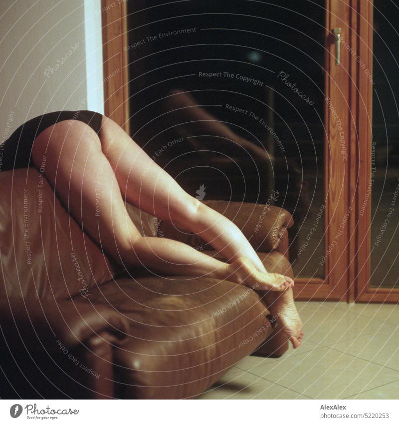 slim, athletic woman in black bodysuit lies on a couch over the backrest and holds her legs in the air in front of a balcony window - analog rectangular photo