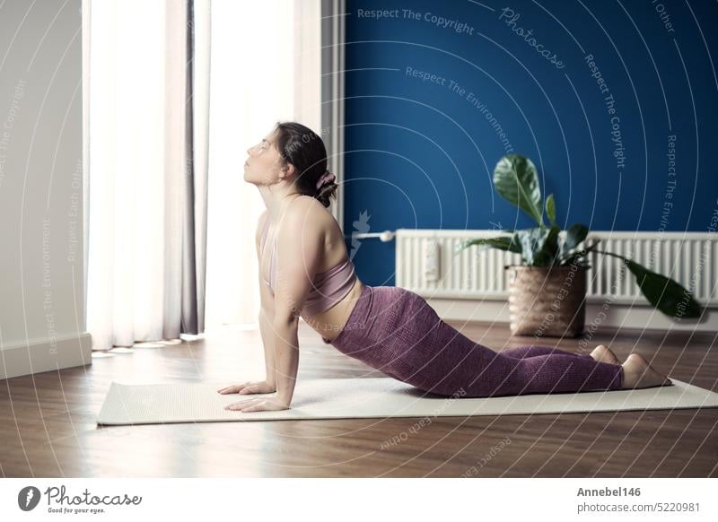 Young sporty woman practicing yoga at home, doing upward facing dog exercise, Urdhva mukha shvanasana pose, working out, wearing sportswear, pants and top, indoor full length, Yoga in living room