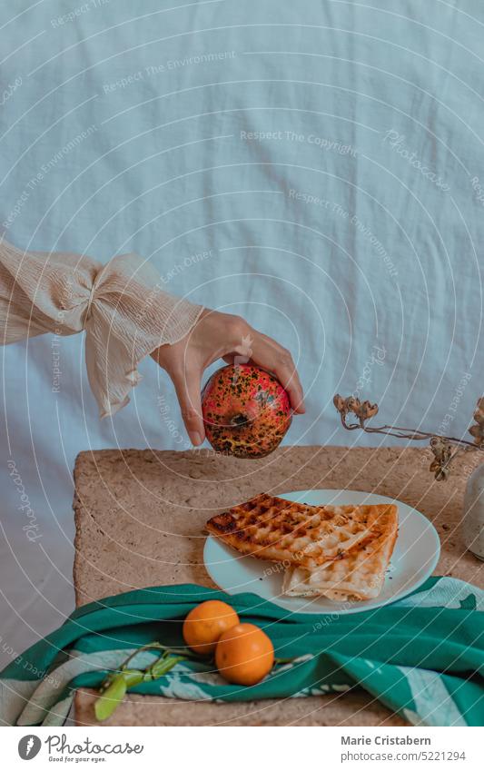Vertical dreamy shot of hand holding a pomegranate over a table with tangerines and waffle for breakfast, concept of springtime, home life, wellness and slow living lifestyle