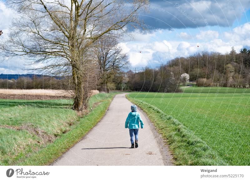 Little child walking on a dirt road through green meadows in spring weather and blue cloudy sky with forest in the distance off the beaten track Nature