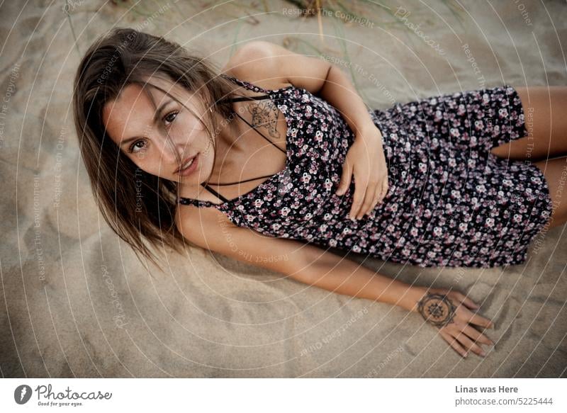 A gorgeous brunette girl is lying down in these sandy dunes. Wearing a summer dress, looking straight into the camera and just being all beautiful. With her pretty face, tanned skin, and inked body.