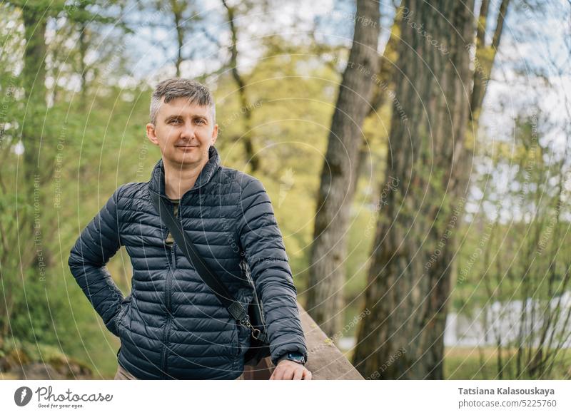 A middle-aged man with a gray hair in a thin down jacket in early spring surrounded by trees forest park nature portrait lifestyle outdoor person caucasian