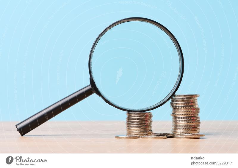 A stack of metal coins and a plastic magnifier on a blue background. The concept of increasing taxes, subsidies magnifying glass money mortgage no people nobody