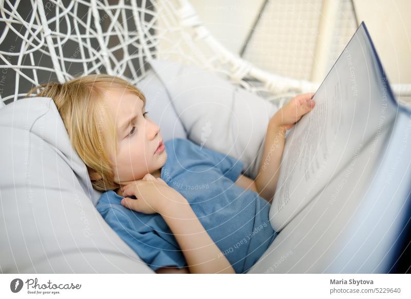 Preteen boy enthusiastically reading book in comfortable chair in balcony. Kid reader enjoying interesting stories, reading fascinating adventurous novels. Child loving to read.
